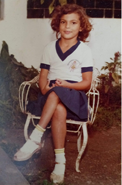 Fotografia antiga de Adriana Cunha quando criança, sentada em uma cadeira branca de ferro, com as pernas cruzadas e as mãos apoiadas no colo. Ela tem cabelos castanhos e cacheados e veste uniforme escolar composto por camiseta branca com detalhes azul-marinho, saia azul, meias brancas e sapatos claros. Ao fundo, há plantas e uma parede clara. Fim da descrição.