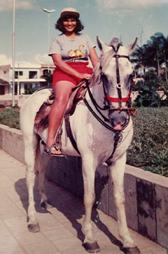 Fotografia antiga de Adriana Cunha sorrindo enquanto monta um cavalo branco. Ela usa boné claro, camiseta cinza com estampa, short vermelho e sandálias. O cavalo está equipado com sela e rédeas. Ao fundo, há uma praça com jardins, palmeiras, construções e pessoas. Fim da descrição.