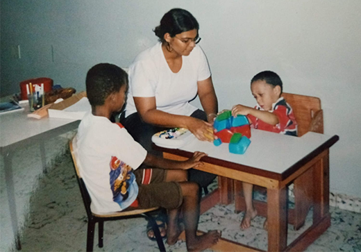 Fotografia da professora Adriana Cunha sentada à mesa com duas crianças em uma atividade com brinquedos coloridos. Ela acompanha a interação dos alunos, dois meninos, enquanto um deles manipula as peças. Ao fundo, há materiais escolares e objetos pedagógicos. Fim da descrição.